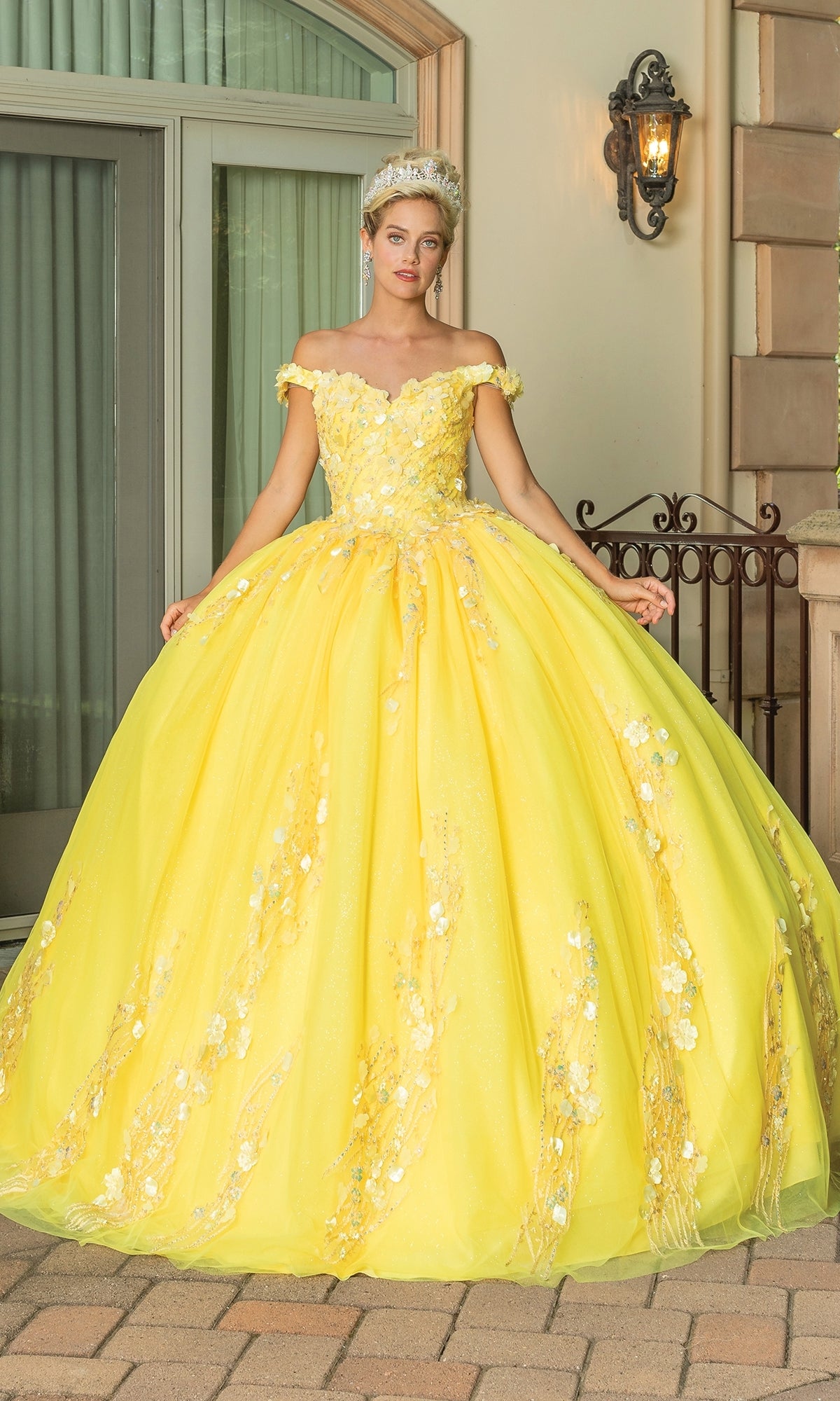 A person stands outdoors on a stone patio wearing the Quinceanera Dress 1775 By Dancing Queen, an off-the-shoulder bright yellow ball gown with floral details and delicate lace embroidery.